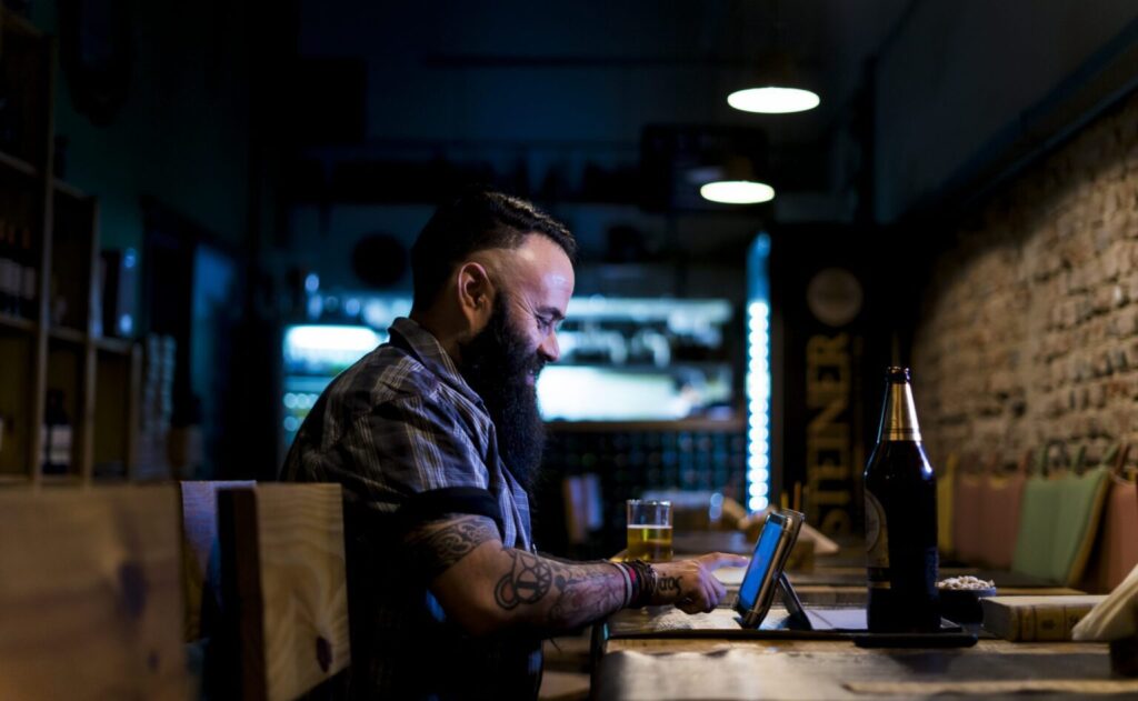 Hombre barbudo usando una tableta en el bar, disfrutando de una cerveza.
