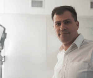 Hombre sonriendo en camisa blanca, frente a pizarra blanca.