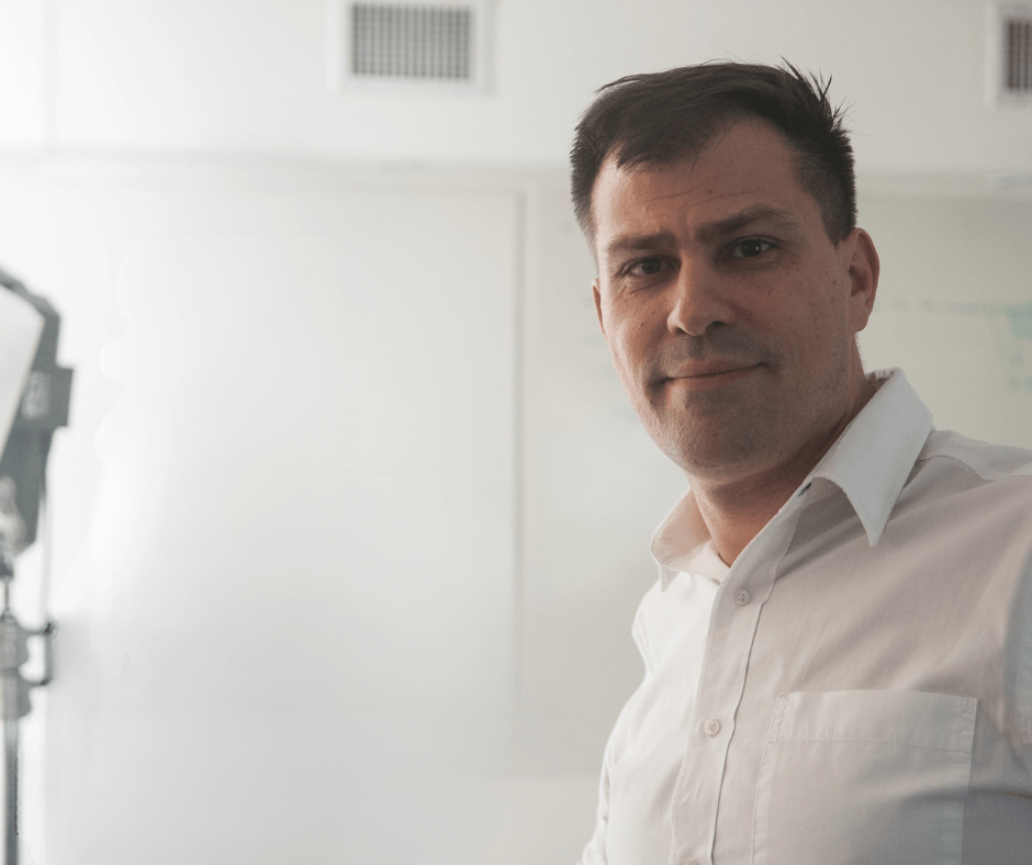 Hombre sonriendo en camisa blanca, frente a pizarra blanca.